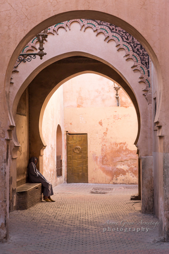 Man wearing traditional Djellaba and Qob (hood) sitting under pink plastered archways outside the former school, Medersa Ben Youssef in the Medina (old walled city), Marrakech.