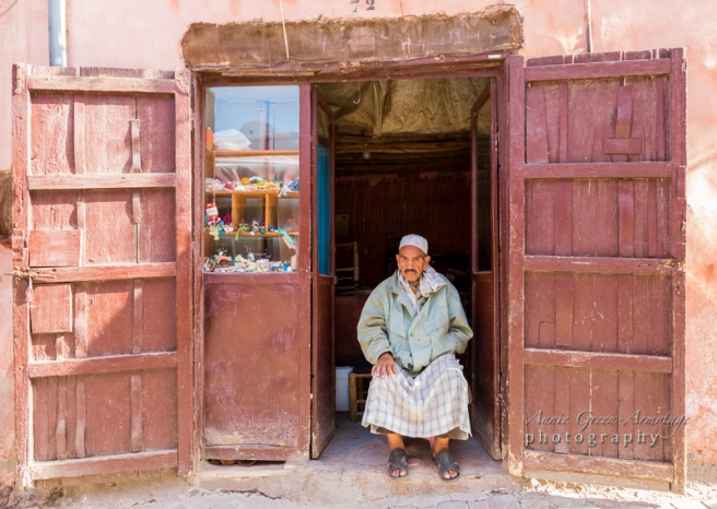 Old man wearing traditional white skullcap and djellaba covered by modern day coat, sits in the doorway of his shop in the souks of the Medina (old walled city), Marrakech. The shop has faded red wooden doors in rough pink plaster walls,and a window with assorted tangled embroidery threads, plus a packet of cigarettes.